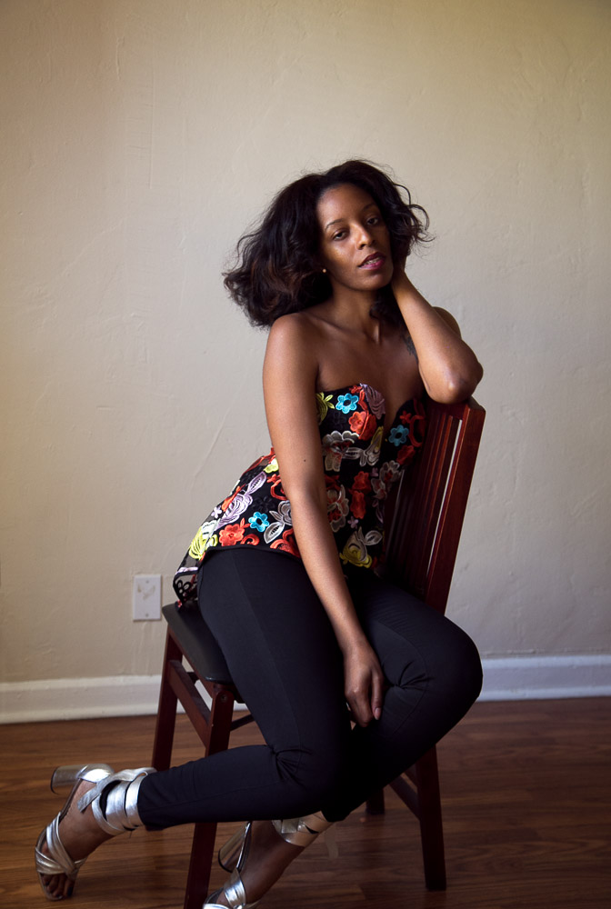 Image is a of a fashionable woman, sitting in a sideways turned chair, looking at the camera.